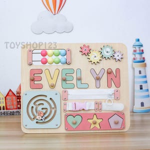 Könnte beinhalten: Holz-Lernspielzeug mit dem Namen "EVELYN" in bunten Buchstaben. Zu den Merkmalen gehören ein Labyrinth, ein Reißverschluss, eine Schnalle, Zahnräder und ein Formensortierer. Das Spielzeug ist für die frühkindliche Entwicklung und das Lernen konzipiert.