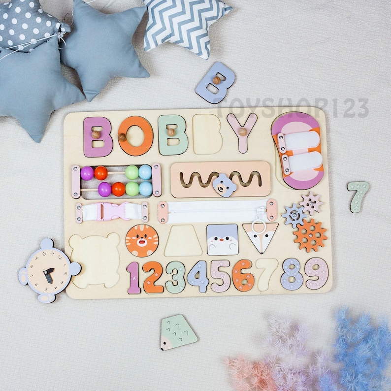 K&ouml;nnte beinhalten: Holz-Lernspielzeug mit dem Namen "BOBBY" in bunten Buchstaben. Das Brett enth&auml;lt einen Abakus, Zahnr&auml;der und die Zahlen 1-9. Zus&auml;tzliche Holzformen und eine Uhr sind ebenfalls enthalten. Ein tolles Lernwerkzeug.