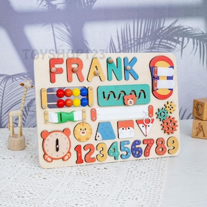 K&ouml;nnte beinhalten: Holz-Lernspielzeug mit dem Namen "FRANK" in bunten Buchstaben. Das Brett enth&auml;lt verschiedene interaktive Elemente, darunter Perlen, einen Rei&szlig;verschluss, Zahnr&auml;der und Tierformen. Zahlen von 1-9 sind ebenfalls enthalten, zusammen mit einem Zifferblatt.