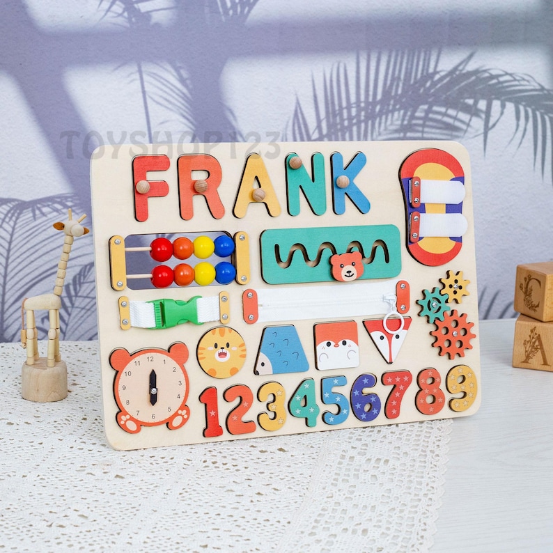 K&ouml;nnte beinhalten: Holz-Lernspielzeug mit dem Namen "FRANK" in bunten Buchstaben. Das Brett enth&auml;lt verschiedene interaktive Elemente, darunter Perlen, einen Rei&szlig;verschluss, Zahnr&auml;der und Tierformen. Zahlen von 1-9 sind ebenfalls enthalten, zusammen mit einem Zifferblatt.