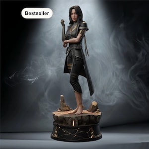 May include: A detailed figurine of a woman in a black and gold outfit, standing on a rocky base. The figurine has a long coat, and the word "Bestseller" is in the upper left corner. The base has a gold geometric design.