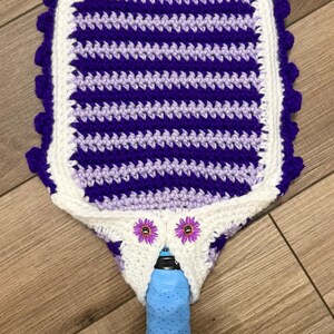May include: A crocheted pickleball paddle cover in white, purple, and lavender. The cover has a striped design and is edged with purple bobbles. Two small flower embellishments are near the handle opening. The paddle handle is blue.