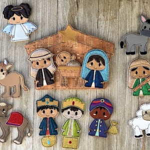 May include: A collection of felt Christmas nativity scene figures, including an angel, Mary, Joseph, baby Jesus, three wise men, a shepherd, a donkey, a cow, a camel, and a lamb. The figures are in various colors, including blue, brown, green, and gold, and are arranged on a wooden surface.