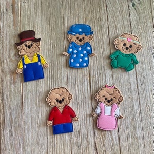 May include: Five felt finger puppets featuring bear-like characters in various outfits. One wears a brown hat and blue overalls, another a blue dress with white polka dots, and a third a green onesie. The other two puppets are wearing red and pink outfits.