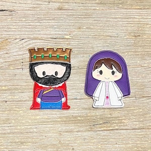 Puede incluir: Dos apliques bordados: un rey con corona dorada, capa roja y túnica morada, y una mujer con tocado morado y túnica blanca. Los apliques están sobre una superficie de madera marrón claro.