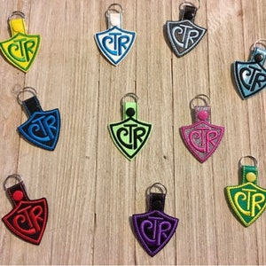 May include: A collection of shield-shaped keychains in various colors, including yellow, blue, green, and purple. Each keychain features the embroidered letters "CTR" in a contrasting color, with a metal keyring attached.