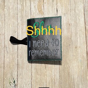 May include: Black notebook cover with the embroidered text "Shhh I need to remember" in green and white. The cover has a black elastic band and green stitching around the edges.