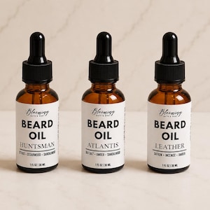 May include: Three amber glass bottles of beard oil with black dropper caps and white labels. Each label reads Blooming Bath & Body, Beard Oil, and the scent names Huntsman, Atlantis, or Leather, with 1 fl oz | 30 ml printed at the bottom.