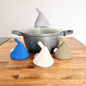 Può includere: Una pentola grigia maculata con coperchio in vetro, circondata da quattro alzacoperchi colorati. Gli alzacoperchi sono blu, bianchi, verdi e grigi. Gli alzacoperchi sono progettati per impedire la tenuta del coperchio.