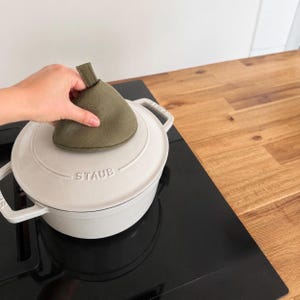 Pode incluir: Uma panela redonda de ferro fundido esmaltado branco com uma tampa apresentando o texto em relevo STAUB. Um pegador de panela triangular de tecido verde-oliva com uma pequena alça está colocado sobre o puxador da tampa.