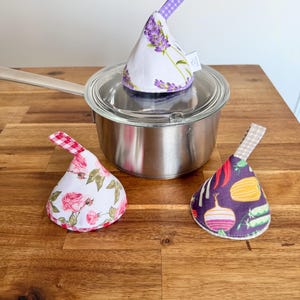 Può includere: Tre presine in tessuto con manici ad anello. Una è su una pentola con coperchio in vetro, con una stampa di lavanda. Le altre due sono accanto alla pentola, una con un motivo a rose e l'altra con una stampa di verdure. Tutte hanno una forma a cono.