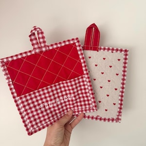 Puede incluir: Dos agarraderas acolchadas. Una es roja con un borde de cuadros blancos y un centro acolchado rojo. La otra es blanquecina con corazones rojos y un borde de cuadros rojos. Ambas tienen un lazo rojo para colgar.
