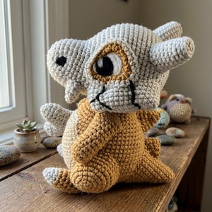May include: A crochet amigurumi creature featuring a light grey skull-like head with black stitched details, large white and black eyes, and a tan-coloured body. The figure has small arms, a short tail, and a textured yarn construction throughout.