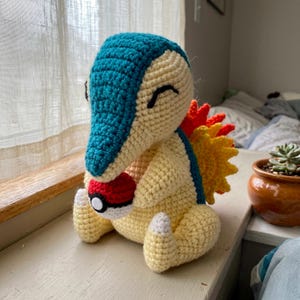Peut inclure: Un jouet en peluche crocheté en forme de créature avec un corps crème, une tête et un dos bleu sarcelle, et une structure en éventail épineuse orange et jaune sur le dos. La créature tient une petite balle ronde rouge, blanche et noire.