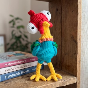 Peut inclure: Un animal en peluche crocheté avec un long cou jaune, un corps bleu sarcelle et des pattes jaunes à trois doigts. La tête est rouge avec deux grands yeux blancs sur des tiges, et un petit col couleur pêche entoure la base du cou.