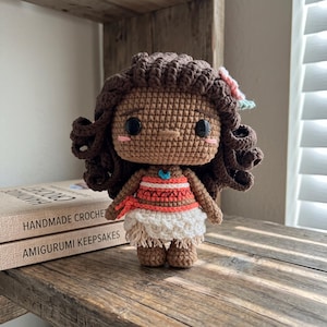 May include: A crocheted doll with dark brown curly hair, a small pink flower accessory, and black bead eyes. The doll wears a coral and white patterned top with a blue pendant and a white skirt with a fringed hem, featuring pink blush on the cheeks.