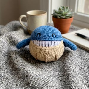 May include: A round, crocheted whale toy featuring a blue upper body and a beige lower body. The whale has a wide, white, stitched mouth with black vertical lines, two small black arched eyes, and two blue side fins extending outward.