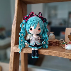 May include: A crocheted doll featuring long, wavy turquoise hair styled in twin tails with black and pink ribbon accents. The doll has a pale face with black eyes, wearing a gray and black outfit with turquoise trim, black arm warmers, and black boots.