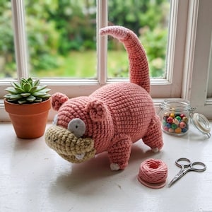 May include: A pink and beige crocheted creature toy with a long curved tail, small ears, and two white teeth. The toy is positioned near a small terracotta pot containing a green succulent, a glass jar filled with colourful beads, a ball of yarn, and scissors.