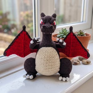May include: A dark brown crocheted dragon plush toy featuring a cream-colored belly, red and black wings, and white claws. The creature has red eyes and a textured yarn construction, standing in an upright position with its arms raised forward.