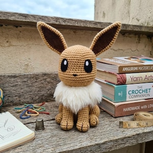 May include: A crocheted Eevee plush toy with light brown yarn, large pointed ears with dark brown centers, black eyes with white highlights, and a fluffy white faux fur collar. The figure sits upright with visible paws and stitched details.