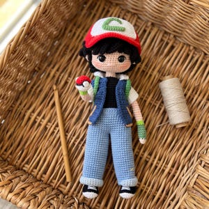 Puede incluir: Una muñeca tejida a mano en crochet que representa un personaje con cabello negro, vistiendo una gorra roja y blanca con un símbolo verde, un chaleco azul sobre una camiseta negra, pantalones azul claro con dobladillo y zapatos blanco y negro.