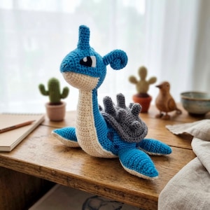 Puede incluir: Un peluche tejido a mano en crochet con forma de criatura marina azul y crema con un caparazón gris en la espalda. La figura presenta un cuello largo, un pequeño cuerno en la cabeza y grandes ojos blancos y negros. Está hecho con puntos visibles.