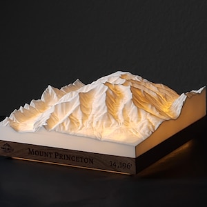 May include: A 3D topographical model of Mount Princeton, with the text "Mount Princeton 14,196'" on a wooden base. The mountain is white and illuminated from within, creating a warm glow. The model is a unique decorative item.