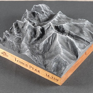 May include: A detailed 3D model of Longs Peak, a mountain with a height of 14,259 feet. The model features a gray, textured surface representing the mountain's terrain, set within a wooden frame. The name "Longs Peak" is engraved on the frame.