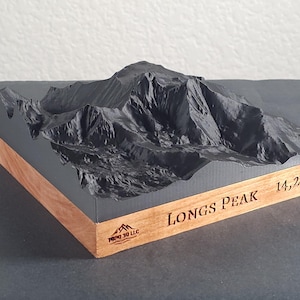 May include: A 3D-printed model of Longs Peak, a mountain in Colorado, USA. The model is dark gray and sits on a wooden base. The mountain's elevation, 14,259', is engraved on the base, along with the name "LONGS PEAK".