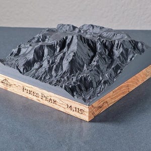 May include: A gray 3D topographical model of Pikes Peak, with the text "PIKES PEAK 14,115" engraved on the wooden base. The mountain model is gray and sits on a wooden base.
