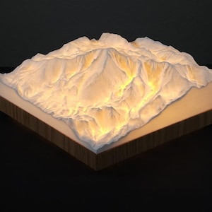 May include: A lighted mountain range sculpture with a detailed, textured surface. The peaks are white, and the base is a warm, glowing light. The base is framed with a dark wood border, creating a striking contrast.