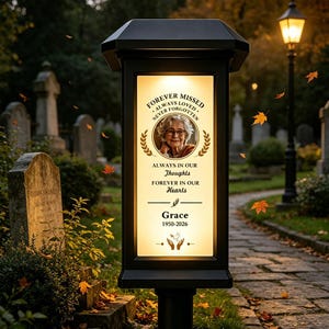 May include: A black solar-powered memorial lantern with a rectangular illuminated panel featuring a portrait of an older person, the text "FOREVER MISSED ALWAYS LOVED NEVER FORGOTTEN", "ALWAYS IN OUR Thoughts FOREVER IN OUR Hearts", "Grace", and "1950-2026".