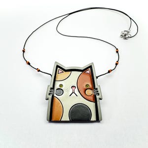 Pode incluir: Um colar com pingente em forma de gato com um cordão preto e fecho prateado. O pingente apresenta um rosto de gato estilizado com manchas laranja, pretas e brancas e olhos verdes. O colar inclui pequenas contas de madeira.