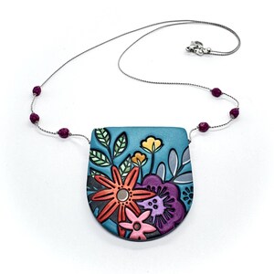 Pode incluir: Colar artesanal com um pendente floral. O pendente turquesa apresenta flores e folhas coloridas. O colar tem uma corrente prateada com pequenas contas redondas de cor vermelho escuro. O pendente tem cerca de 4 cm de largura.