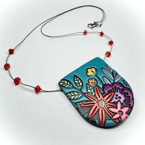 Pode incluir: Um colar artesanal com um pingente turquesa com um design floral. O pingente apresenta flores laranja, rosa, amarelas e roxas com folhas verdes. O colar inclui uma corrente prateada e contas vermelhas.