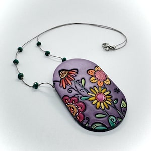 Pode incluir: Colar artesanal com um pingente lavanda com um design floral em tons de laranja, amarelo e rosa. A corrente prateada tem contas verdes. O pingente tem cerca de 5 cm de comprimento, uma joia única.
