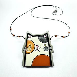 Pode incluir: Um colar com pingente em forma de gato com um cordão preto e fecho prateado. O pingente apresenta um design de gato branco, laranja e preto com uma borda cinza. Pequenas contas laranja adornam o cordão perto do pingente.