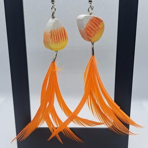 May include: A pair of dangle earrings with a candy corn design. The earrings feature white, orange, and yellow painted teardrop-shaped pendants, with long orange feathers hanging below. The earrings are displayed on a black stand.