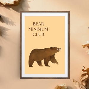 Puede incluir: Un cuadro enmarcado con una ilustración de un oso pardo sobre un fondo amarillo claro. El texto "BEAR MINIMUM CLUB" se muestra encima del oso. La obra de arte está enmarcada en un marco marrón, ideal para la decoración del hogar.