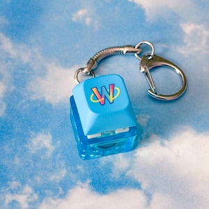 May include: A light blue keycap keychain with a silver-colored clasp. The keycap has a colorful "W" logo. The keychain is attached to a clear, rectangular base. The background is a blue sky with white clouds.