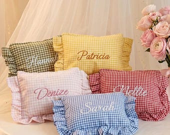 Puede incluir: Cinco cojines rectangulares de cuadros vichy con bordes de volantes, cada uno bordado con un nombre: Hannah, Patricia, Denize, Nellie y Sarah. Los cojines son de color verde, amarillo, rosa claro, rojo y azul, junto a un ramo de rosas.