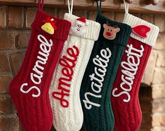 Puede incluir: Cuatro calcetines navideños de punto trenzado colgados en fila. El primero es rojo con el texto blanco Samuel y una campana amarilla. El segundo es blanco con el texto rojo Amelie y una cara de Santa. El tercero es verde con el texto blanco Natalie y una cara de reno. El cuarto es rojo con el texto blanco Scarlett y un gorro rojo.