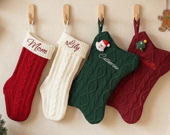 Puede incluir: Cuatro calcetines navideños de punto trenzado colgados en ganchos de madera. El primero es rojo con 'Mom' bordado, el segundo blanco con 'Lily', el tercero verde oscuro con 'Catherine' y un parche de Santa, y el cuarto granate con 'Isabella'.
