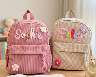 Puede incluir: Dos mochilas de pana, una rosa empolvado y otra beige, cada una con un asa superior y un bolsillo frontal con cremallera. La mochila rosa muestra el nombre Sophie en letras de hilo de colores con detalles florales, mientras que la beige presenta el nombre Finley en letras de hilo similares con flores.