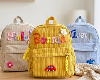 Puede incluir: Tres mochilas de pana en beige, amarillo y azul claro. Cada bolsa tiene un asa superior, un bolsillo frontal con cremallera y un nombre bordado con hilo de colores. La mochila amarilla muestra un coche rojo, mientras que las otras tienen flores.