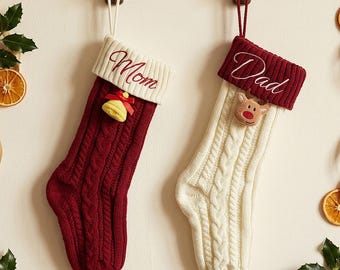 Puede incluir: Dos calcetines navideños de punto trenzado colgados de ganchos de madera. El calcetín izquierdo es rojo oscuro con un puño blanco bordado con la palabra Mom y un adorno de campana amarilla. El derecho es blanco con puño rojo, bordado Dad y un reno.