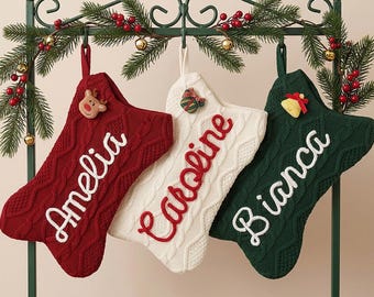 Puede incluir: Tres calcetines navideños de punto trenzado en rojo, blanco y verde oscuro, colgados de un soporte de metal verde decorado con ramas de pino y bayas rojas. Los calcetines presentan los nombres bordados Amelia, Caroline y Bianca en cursiva.