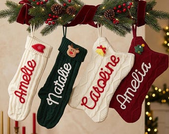 Puede incluir: Cuatro calcetines navideños de punto colgados de una rama de abeto horizontal decorada con bayas rojas, piñas y una cinta de terciopelo rojo oscuro. Los calcetines son blancos, verde oscuro, blancos y rojo oscuro, con los nombres Amelie, Natalie, Caroline y Amelia.