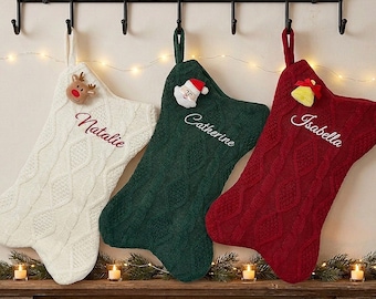 Puede incluir: Tres calcetines navideños de punto trenzado en blanco, verde oscuro y rojo intenso, cada uno con un lazo para colgar. El calcetín blanco tiene un reno, el verde una cara de Papá Noel y el rojo una campana. Los nombres Natalie, Catherine e Isabella están bordados.